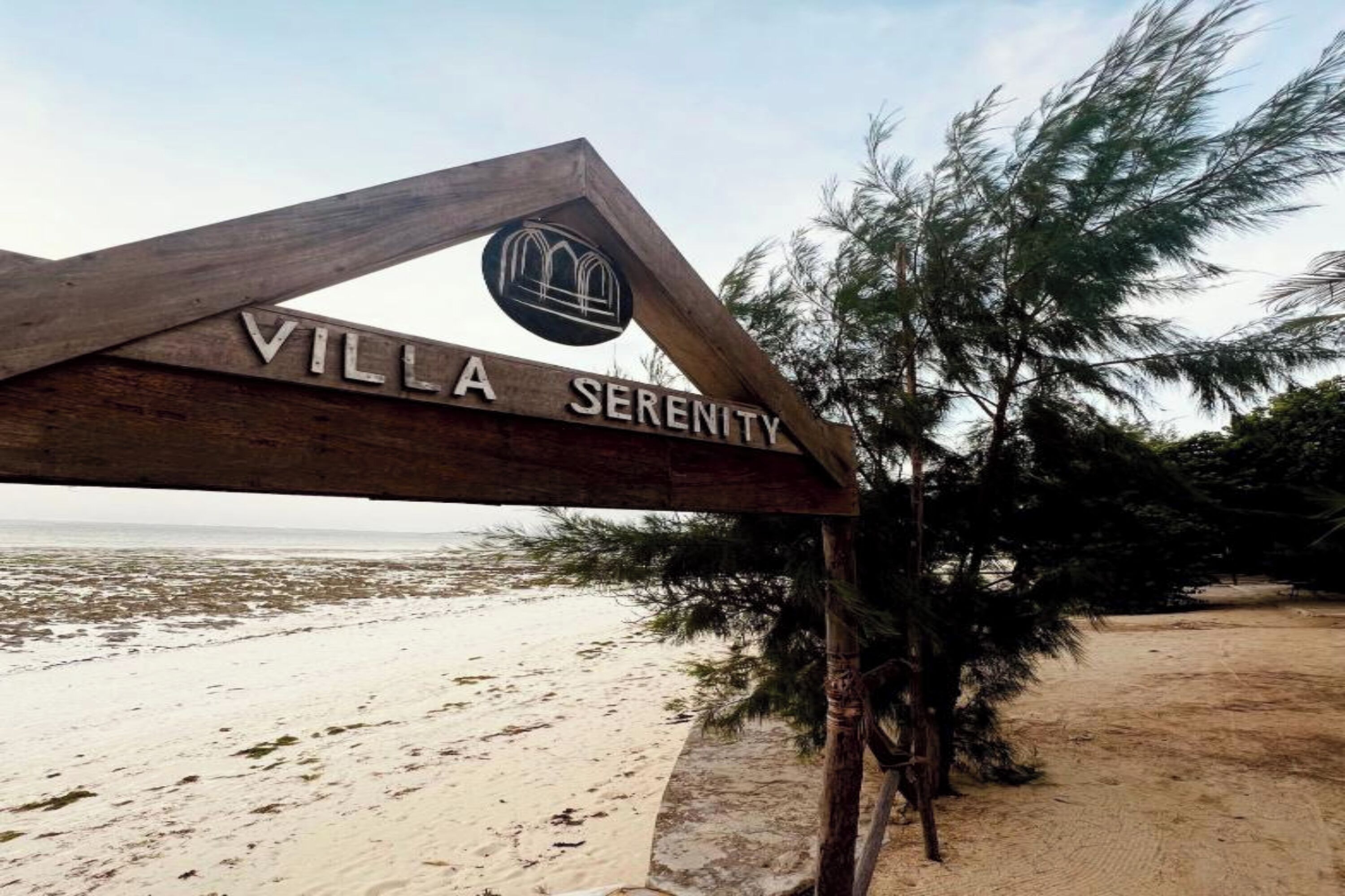 Uma placa de madeira com o texto 'VILLA SERENITY' e um logotipo, numa praia com árvores e areia.