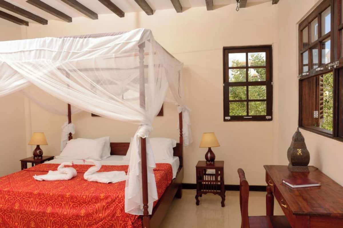A four-poster bed with a canopy, a desk with a lamp, and a window with a view of greenery.