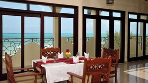 A dining area with a table set for four, large windows offering a view of the ocean, and a balcony with a railing. - Villa Serenity Zanzibar (Paje)
