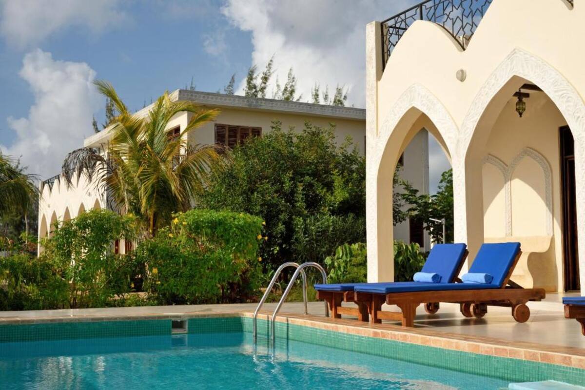 A swimming pool with lounge chairs, surrounded by greenery and a building with arches.