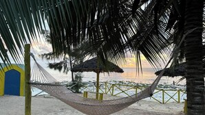 A hammock tied between palm trees on a sandy beach with a thatched-roof structure in the background. - Villa Serenity Zanzibar (Paje)