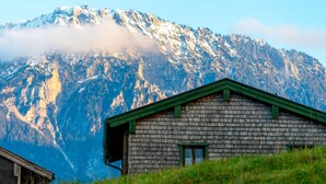 Une cabane en bois rustique, surmontée d’un toit vert, se dressant sur un fond de montagnes enneigées et d’un ciel dégagé.