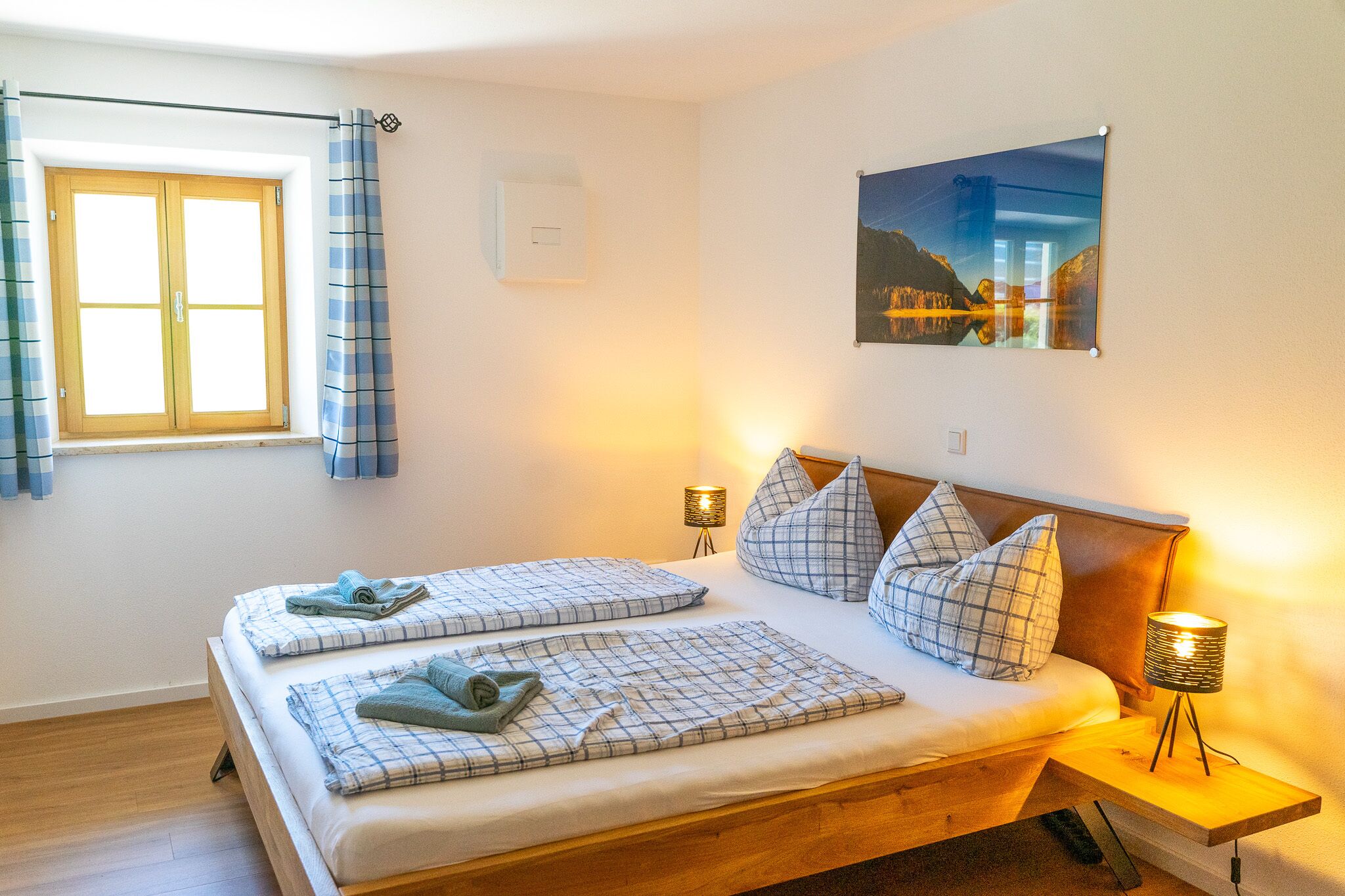 Un lit double avec une tête de lit en bois, deux tables de chevet avec des lampes, une fenêtre avec des rideaux et un tableau au mur.