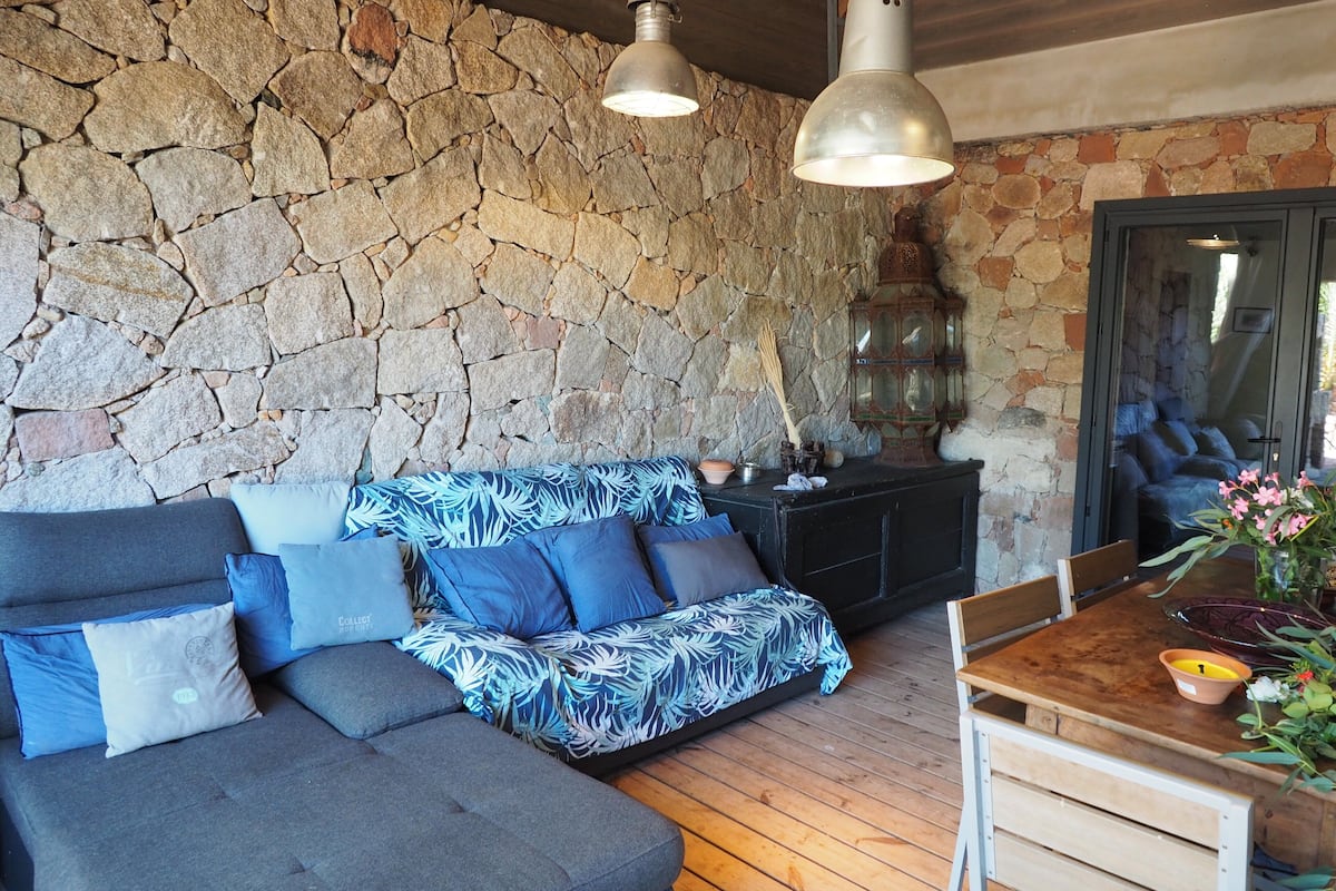 Uma sala de estar aconchegante com parede de pedra, sofá modular com almofadas estampadas, mesa de jantar de madeira e luminárias pendentes.