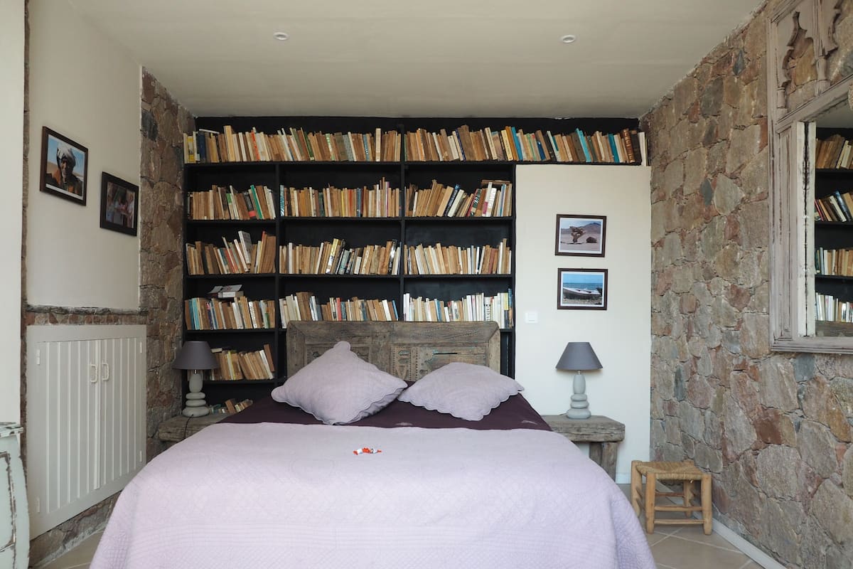 Quarto com uma cama grande, uma estante cheia de livros, uma mesinha com abajur e uma parede de pedra.