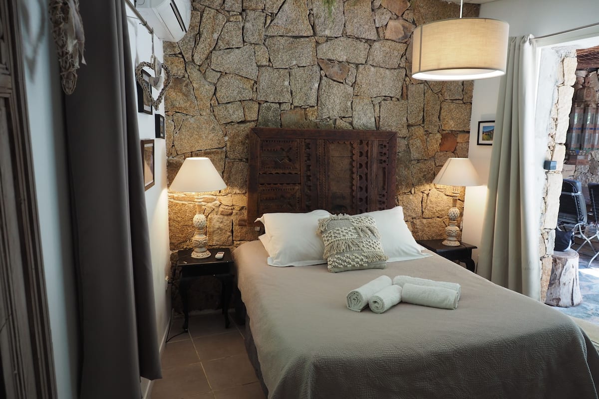 Quarto com parede de pedra, cabeceira de madeira, duas mesas de cabeceira com luminárias, cama com cobertor bege e toalhas dobradas, e luminária pendente.