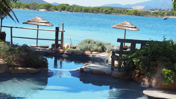 A clear blue pool with a wooden deck and thatched umbrellas, surrounded by rocks and greenery. - Maranatha Porto Vecchio (Porto-Vecchio)