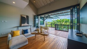 Amplia sala de estar con piso de madera, un sofá gris con cojines amarillos y azules, una mesa de centro y un ventanal con vistas a un follaje exuberante.