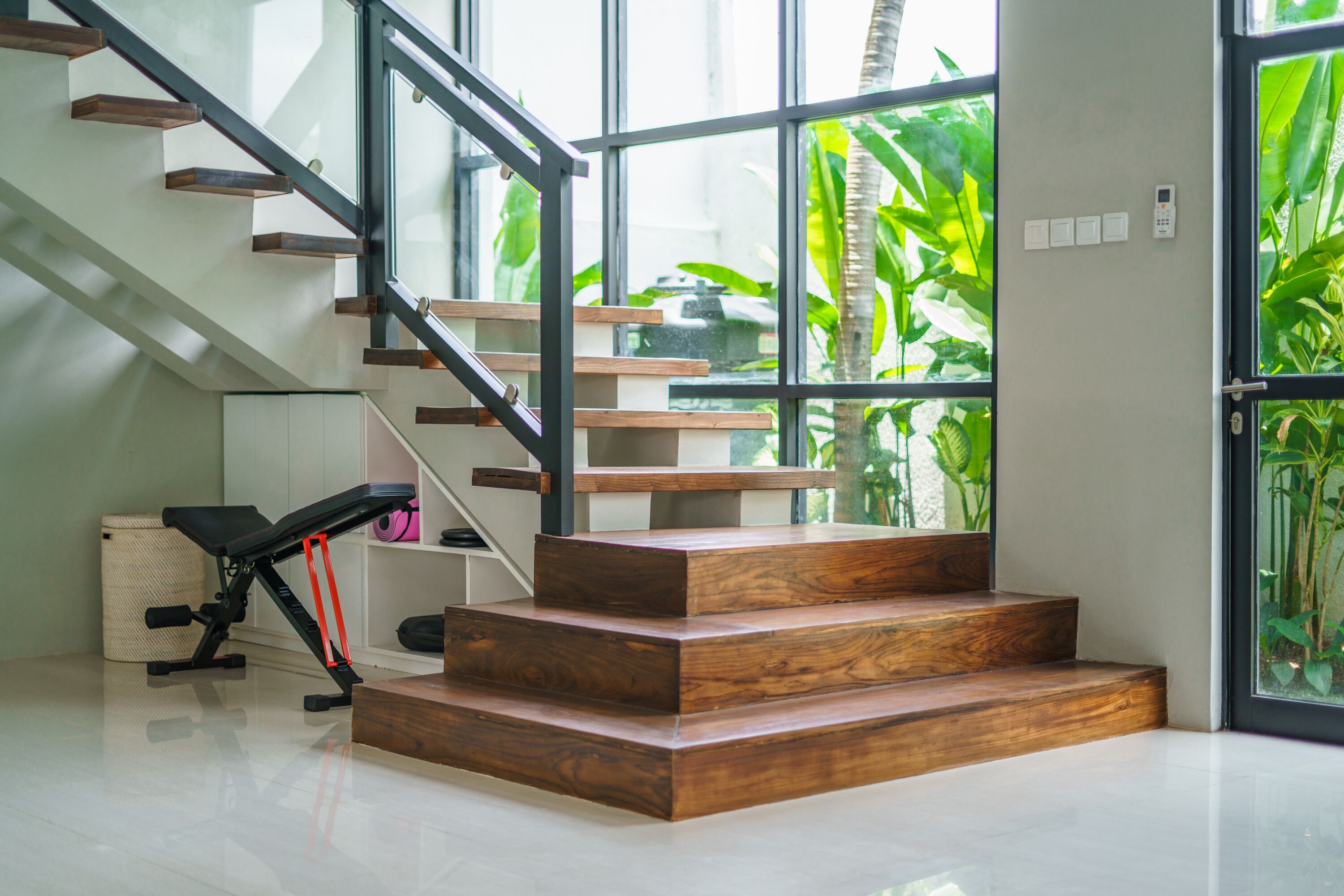 A modern staircase with wooden steps and glass railings, leading to a room with a view of lush greenery outside.