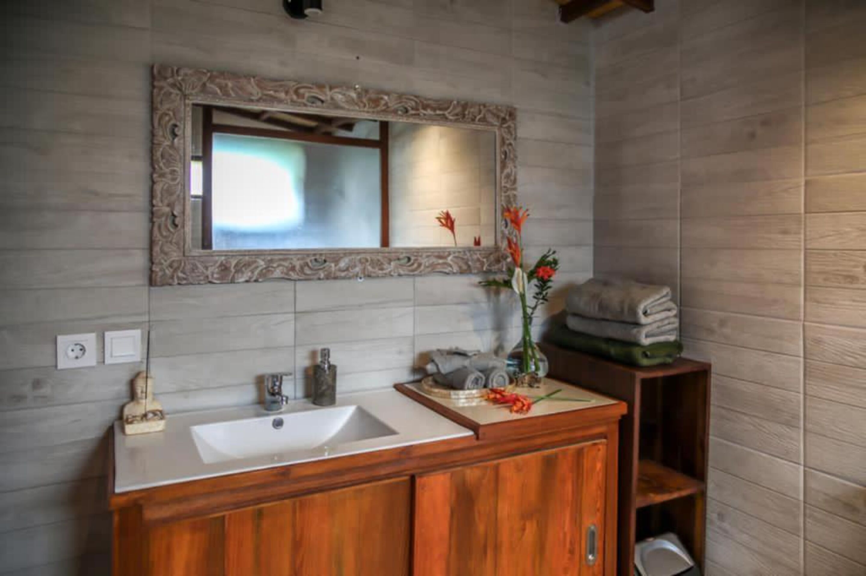 Un baño con un tocador de madera, un espejo grande y un lavabo. Hay una repisa con toallas y un florero con flores.