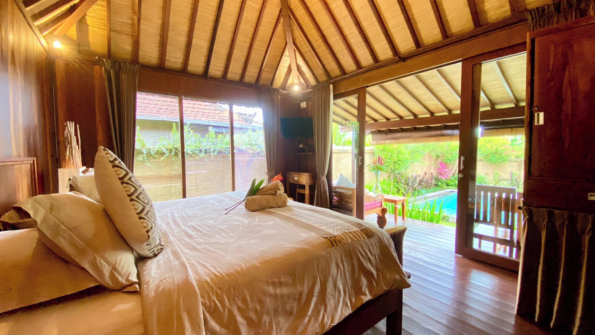 A bedroom with a large bed, a wooden ceiling, and a view of a garden and pool.