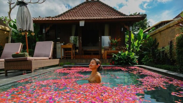 A pool with floating flower petals, lounge chairs, and a traditional building in the background. - NILUH Private Villas  (Ubud)