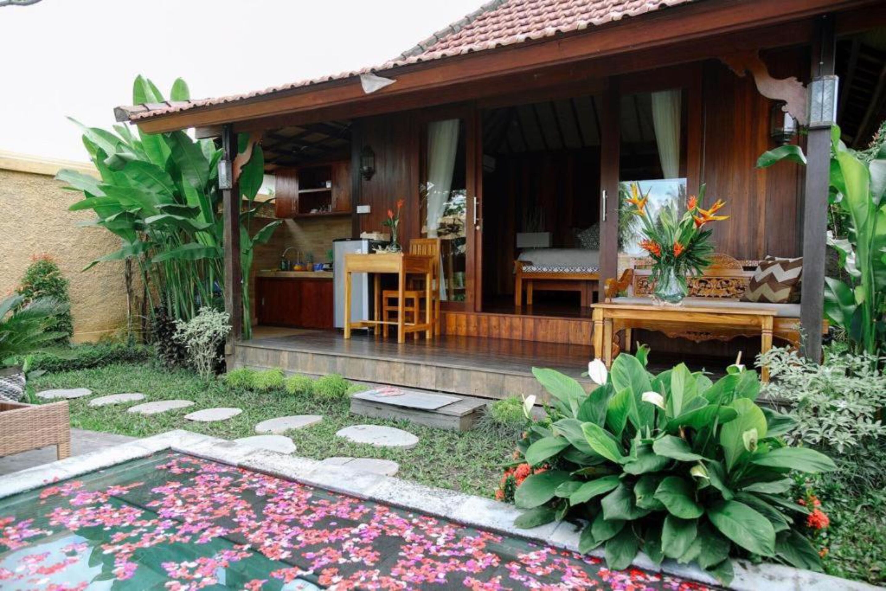 Uma casa de madeira com varanda coberta, área de jantar e uma piscina pequena com flores flutuando.