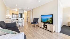 A modern living room with a flat-screen TV, a sofa, a dining area with a white table and chairs, and a kitchen in the background.