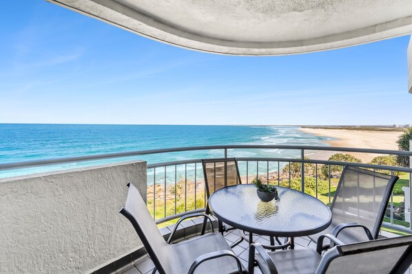 A balcony with a table and chairs overlooking a beach and ocean.