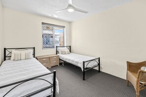 A bedroom with two single beds, a small wooden dresser, a wicker chair, and a window with a view of buildings.