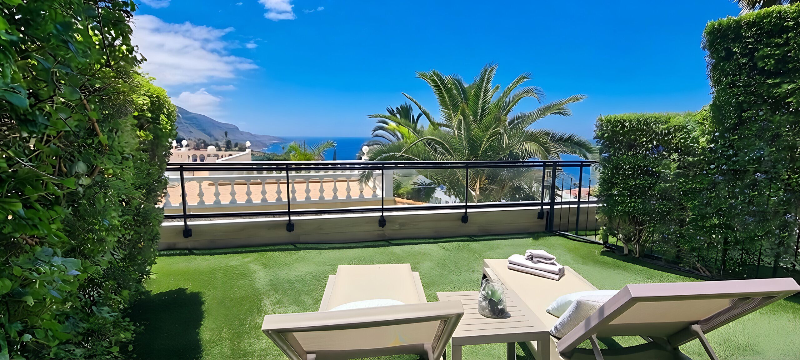 Un balcón con vista al mar, césped verde y mobiliario exterior.