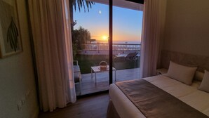 A hotel room with a bed, a view of the sunset, and a balcony with outdoor furniture.