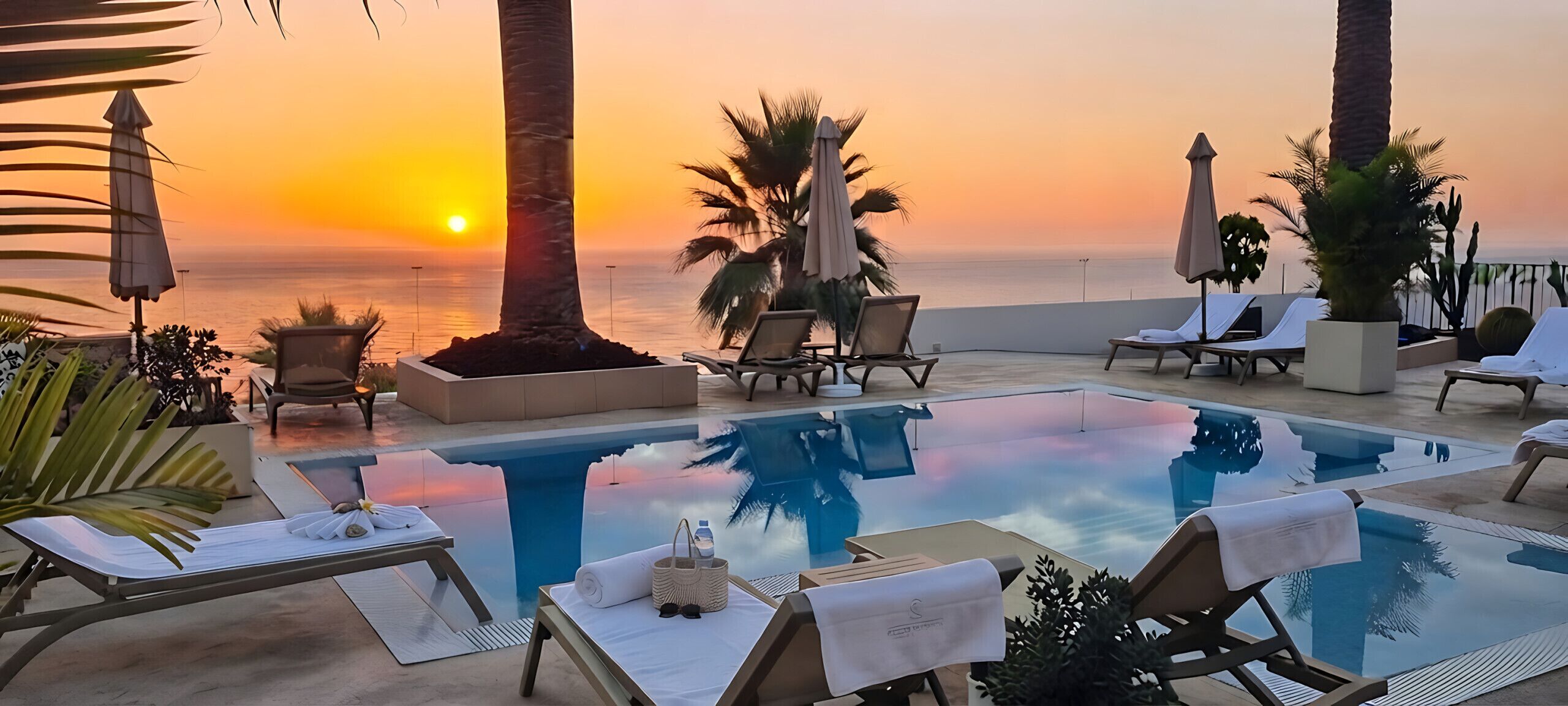 Ein Poolbereich mit Liegestühlen und Sonnenschirmen mit Blick auf einen Sonnenuntergang über dem Meer.