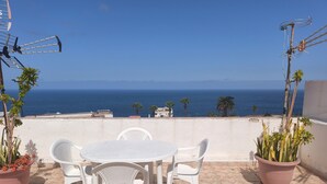 Une terrasse avec une table et des chaises blanches, des plantes en pot et une vue sur l’océan.