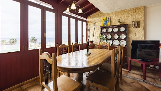 Un coin repas avec une table et des chaises en bois, un lustre, un mur en pierre agrémenté d’une étagère à assiettes et un téléviseur posé sur un meuble.