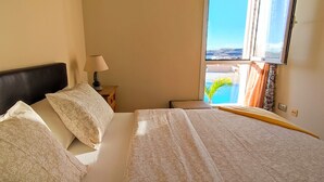 A hotel room with a bed, a nightstand with a lamp, and a view of a pool and mountains through the window. - Coast, 4 people, sea and pool view, free WiFi  (Porís de Abona, Santa Cruz de Tenerife)