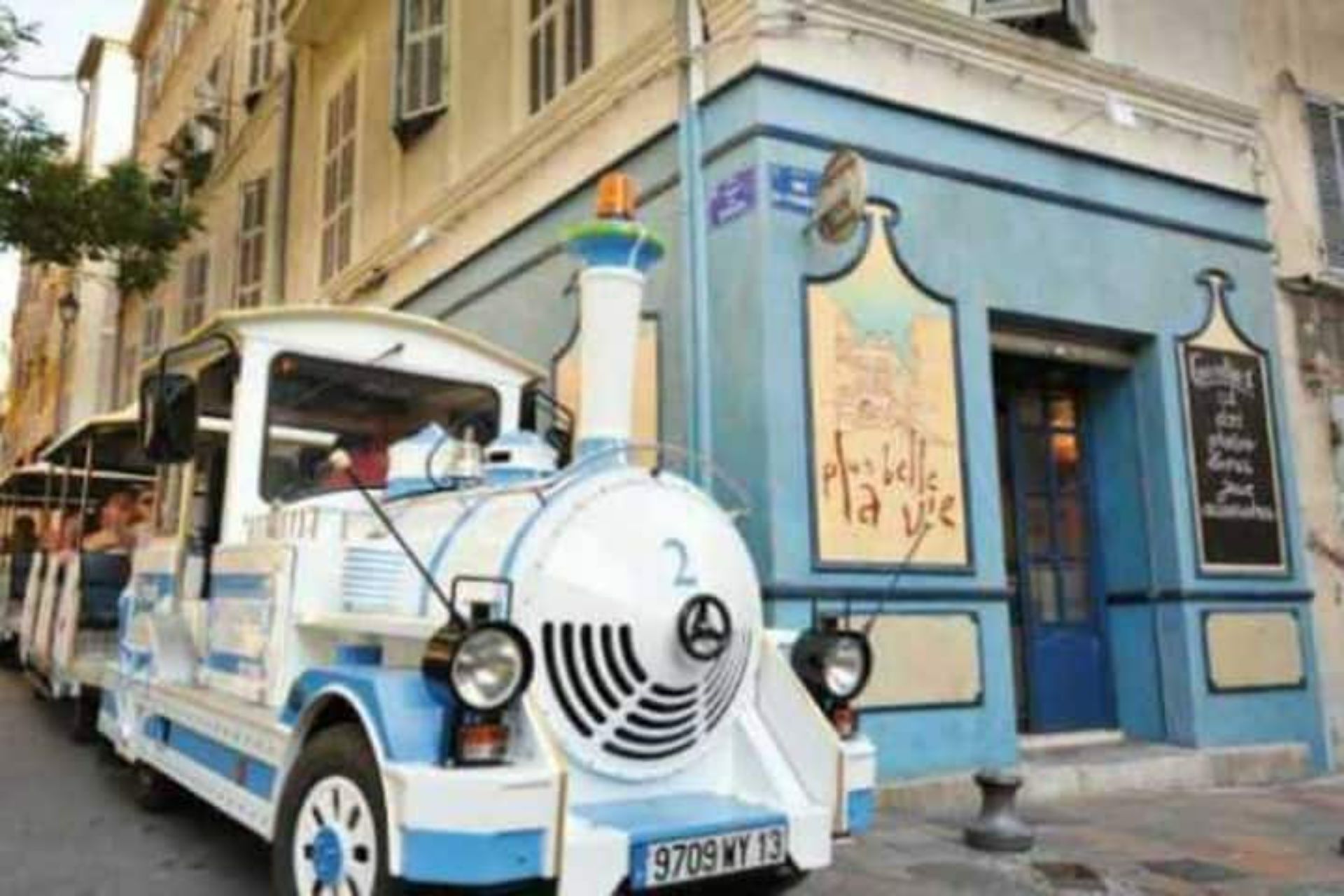 Une voiture ancienne, bleue et blanche, est garée devant un bâtiment sur lequel on peut lire l’inscription « Plas d’Is ».