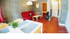 A compact living space with a red sofa, a small table, and a lamp. A plant is on the table, and there's a doorway leading to another room. - Charmant T2 34m2, 2e Étage au Coeur du Panier (Marseille)