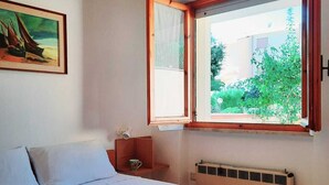 A bedroom with a bed, a window with curtains, a radiator, a small table with a cup, and a painting on the wall. - Baia Asinara - Appartamento Fronte Mare Castelsardo (Lu Bagnu)