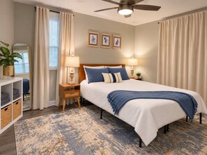 Une chambre à coucher comprenant un lit, des tables de chevet, un ventilateur de plafond, une commode, une lampe, un miroir, une étagère avec des paniers et une fenêtre avec des rideaux.