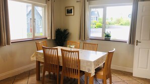 Ein Essbereich mit Tisch und Stühlen, großen Fenstern mit Vorhängen, einer Zimmerpflanze und einer Tür.
