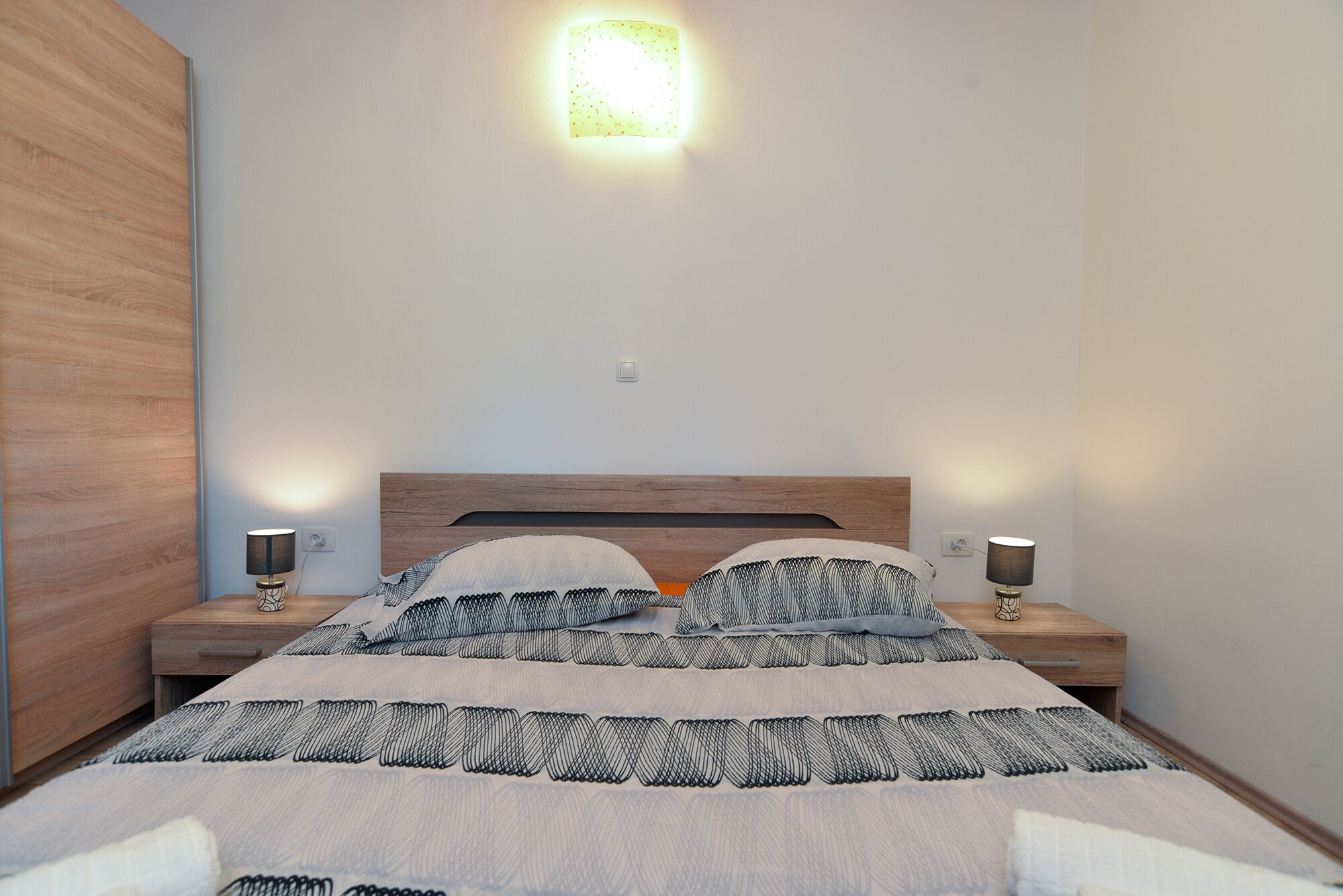 A bedroom with a wooden headboard, two bedside lamps, a patterned bedspread, and a wall-mounted light fixture.