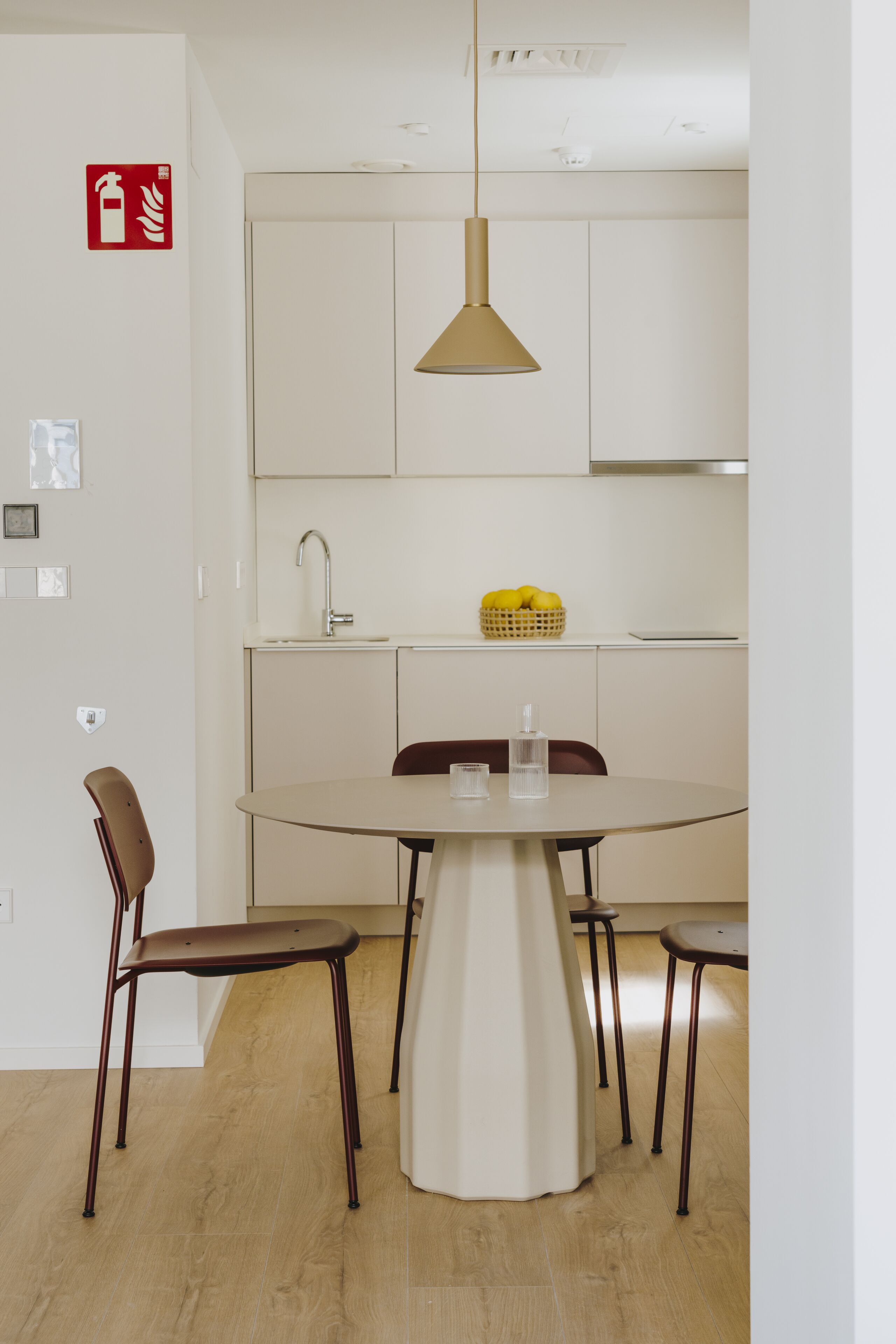 Una cocina moderna con una mesa redonda, dos sillas, una lámpara colgante y una cesta de fruta en la encimera.