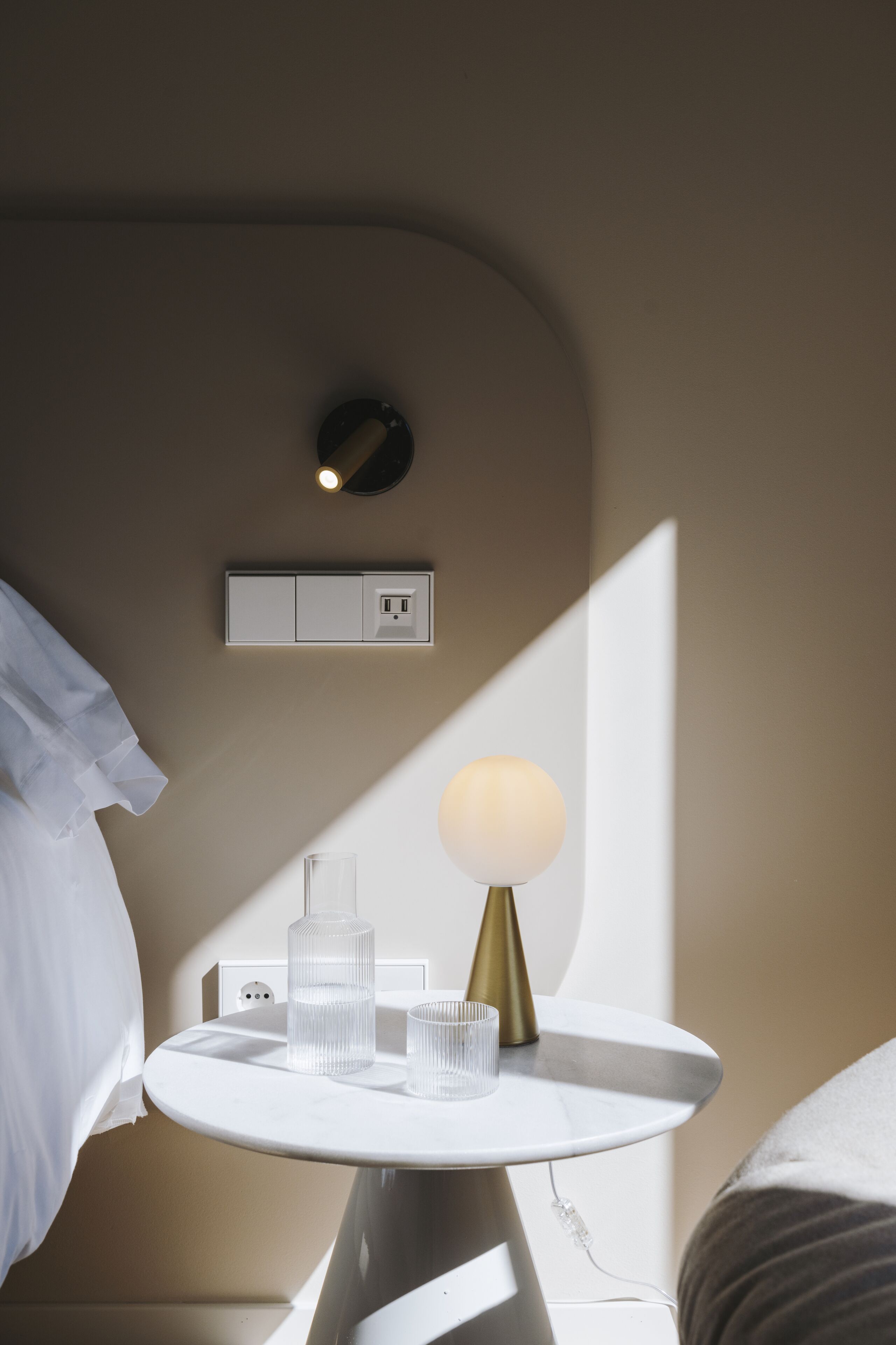 A neatly arranged hotel room with a bedside table featuring a lamp, a glass, and a bottle. A power outlet and a switchboard are visible on the wall.