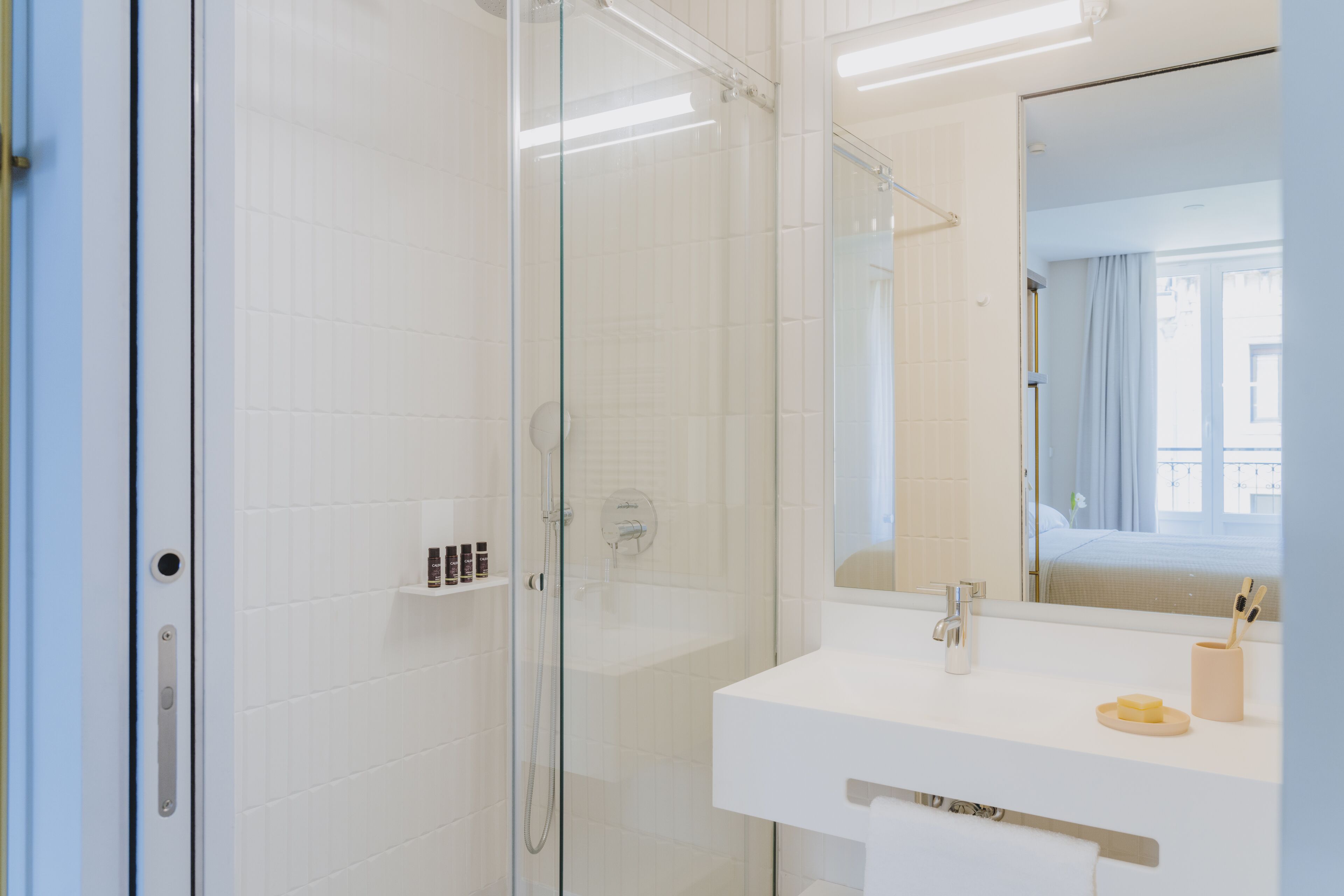 A modern bathroom with a glass shower enclosure, a white sink, and a mirror reflecting a bedroom.