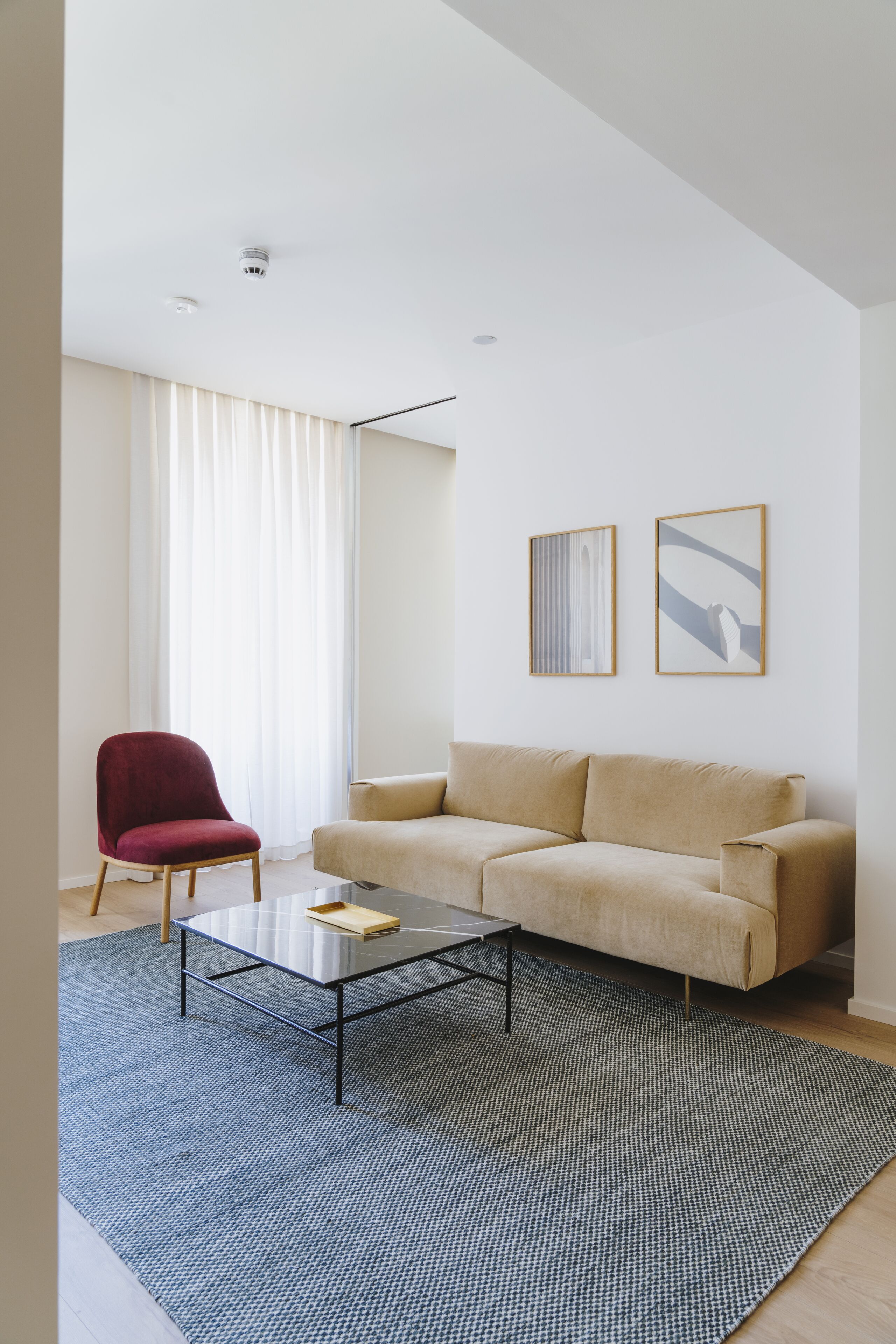 Ein modernes Wohnzimmer mit einem beigefarbenen Sofa, einem roten Sessel und einem Glastisch. An der Wand befinden sich zwei gerahmte Kunstwerke und ein Fenster mit weißen Vorhängen.