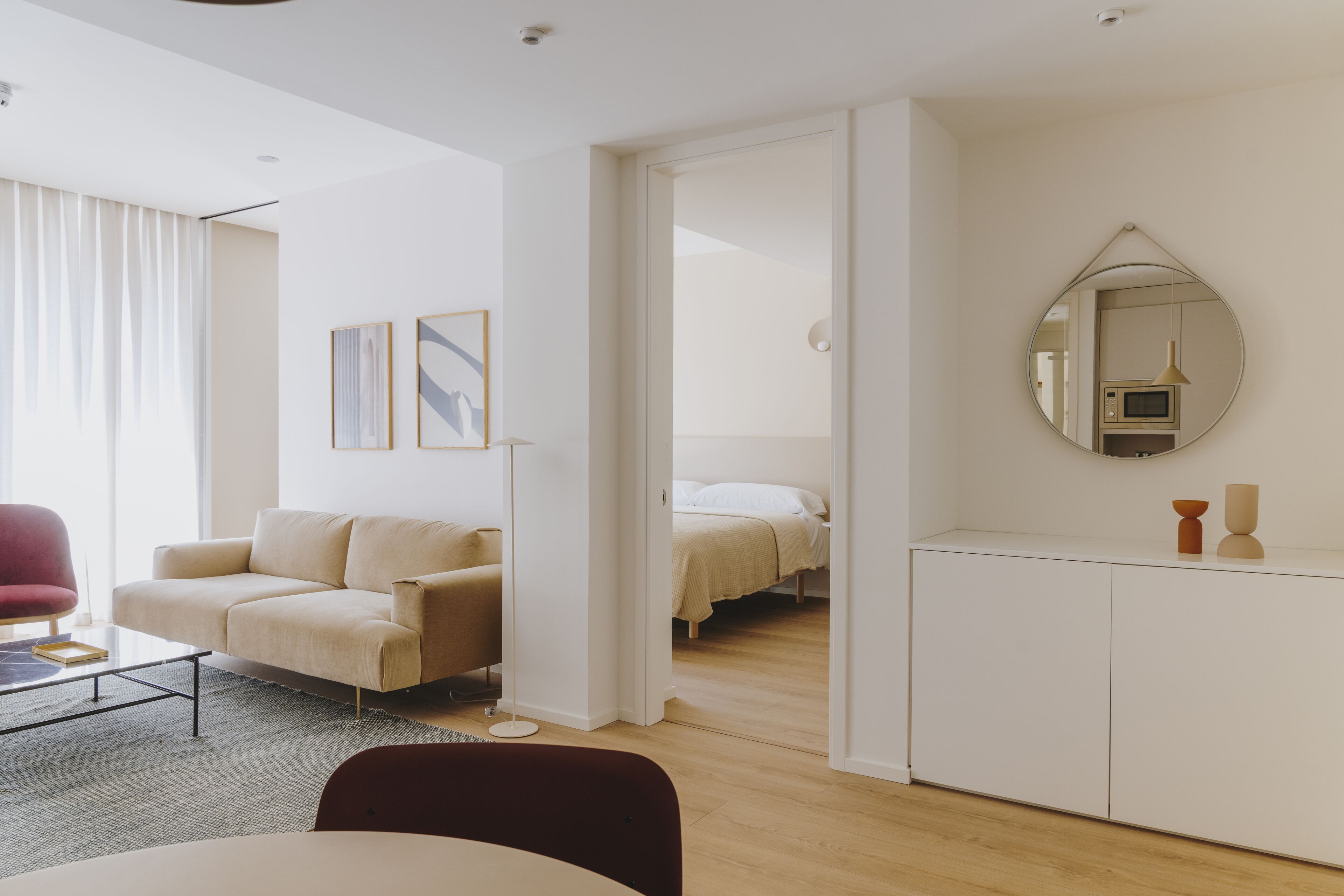 Una sala de estar moderna y bien iluminada con un sofá, una mesa de centro y un comedor. Hay una cama en la habitación contigua y un espejo en la pared.