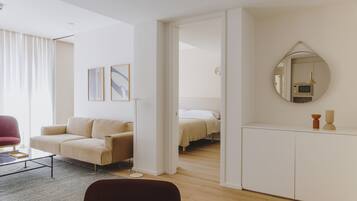 Una sala de estar moderna y bien iluminada con un sofá, una mesa de centro y un comedor. Hay una cama en la habitación contigua y un espejo en la pared.