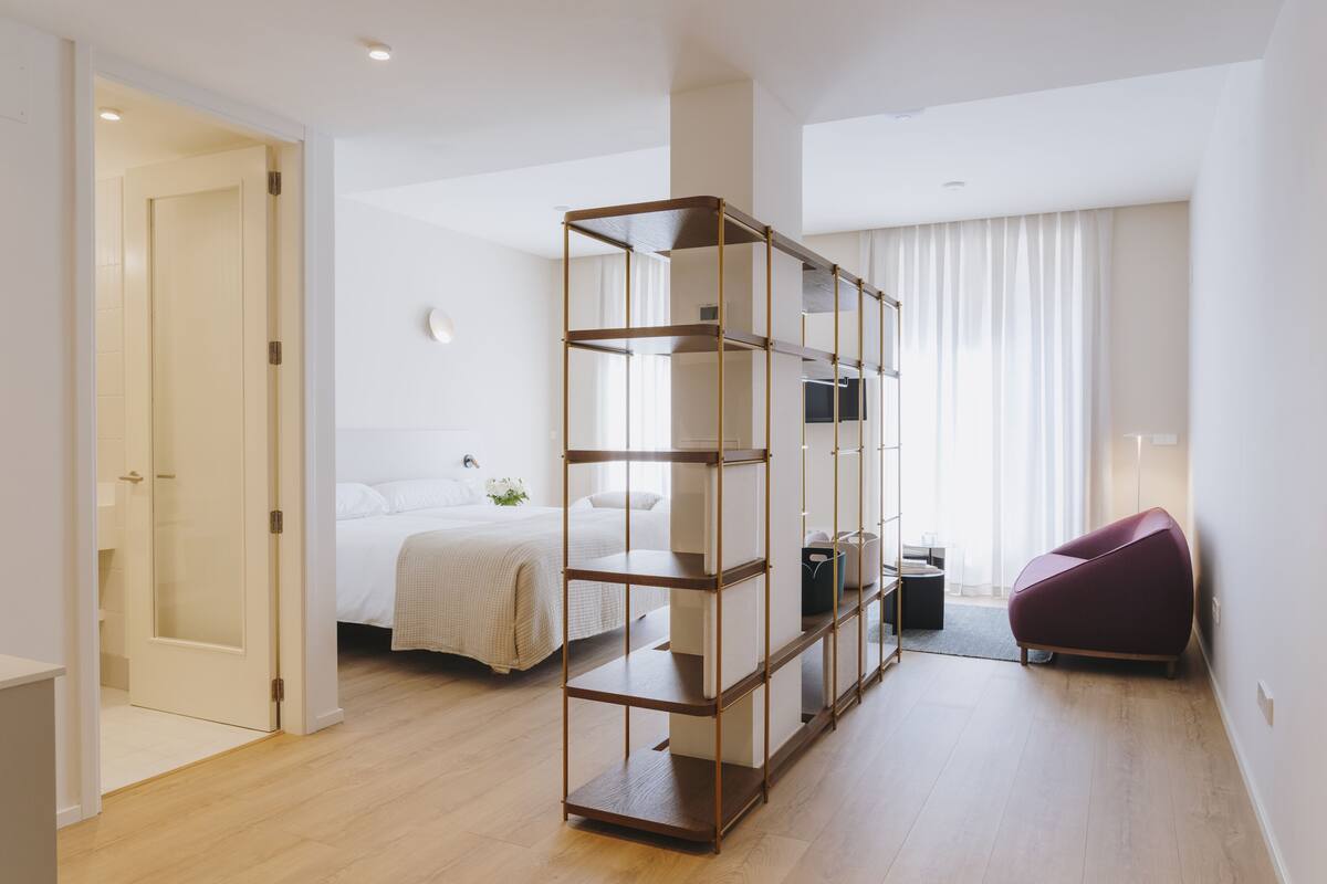 A modern hotel room with a wooden floor, a bed with white bedding, a red chair, a metal shelving unit, and a large window with curtains.