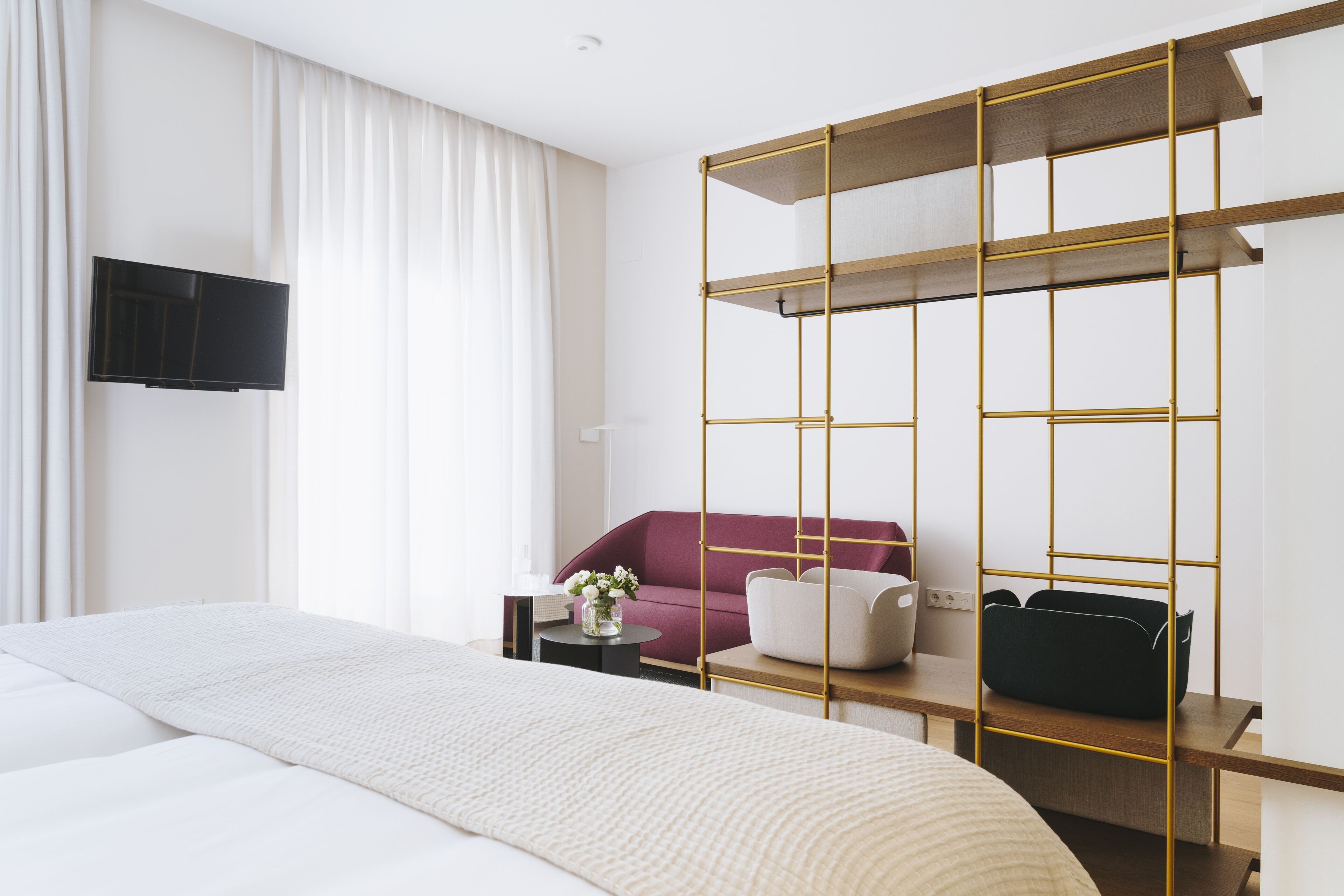 Une chambre d’hôtel moderne avec un grand lit, un téléviseur fixé au mur, un canapé rouge et une étagère en bois.