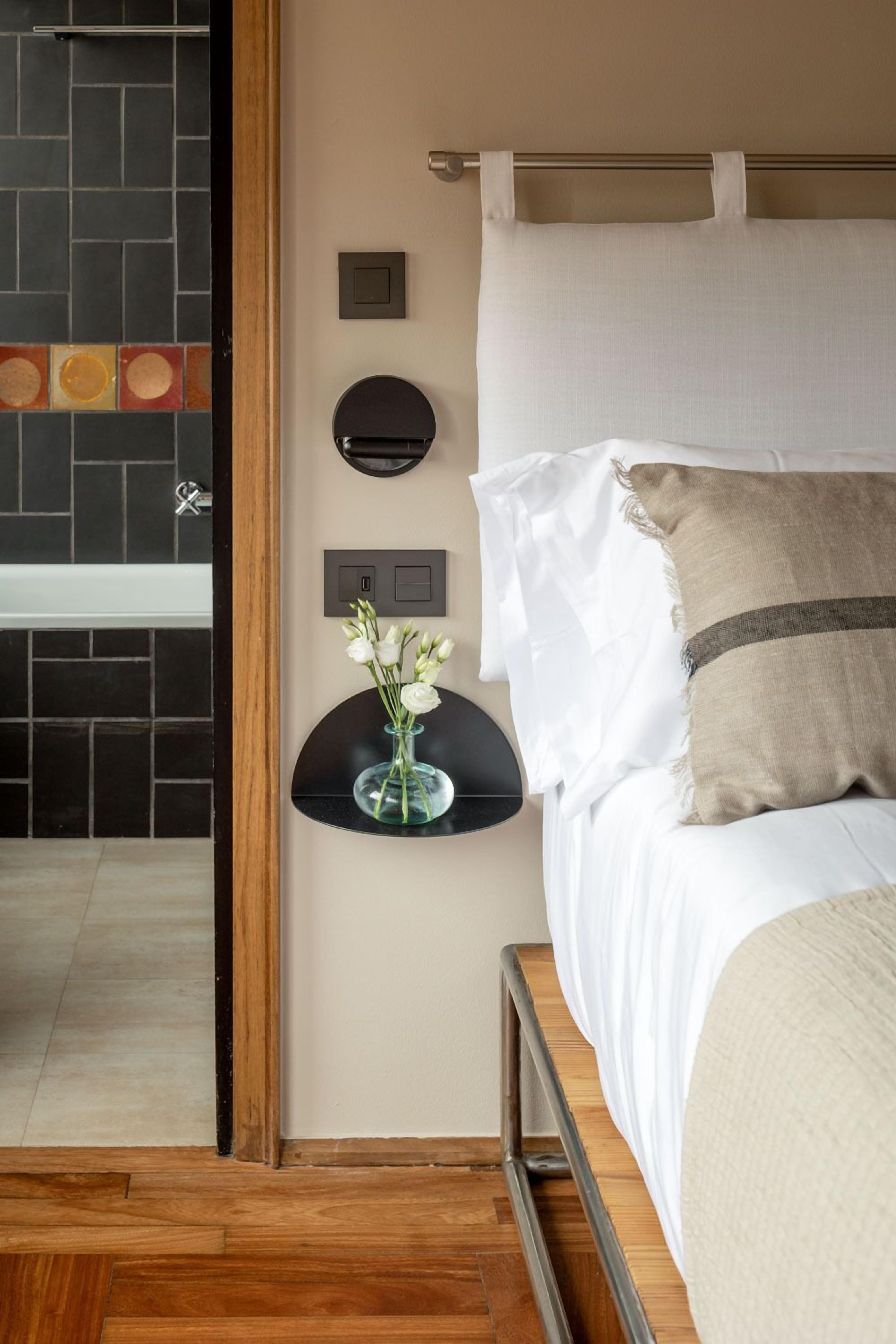 Un dormitorio con cama, mesita de noche con un florero y una lámpara fijada a la pared.