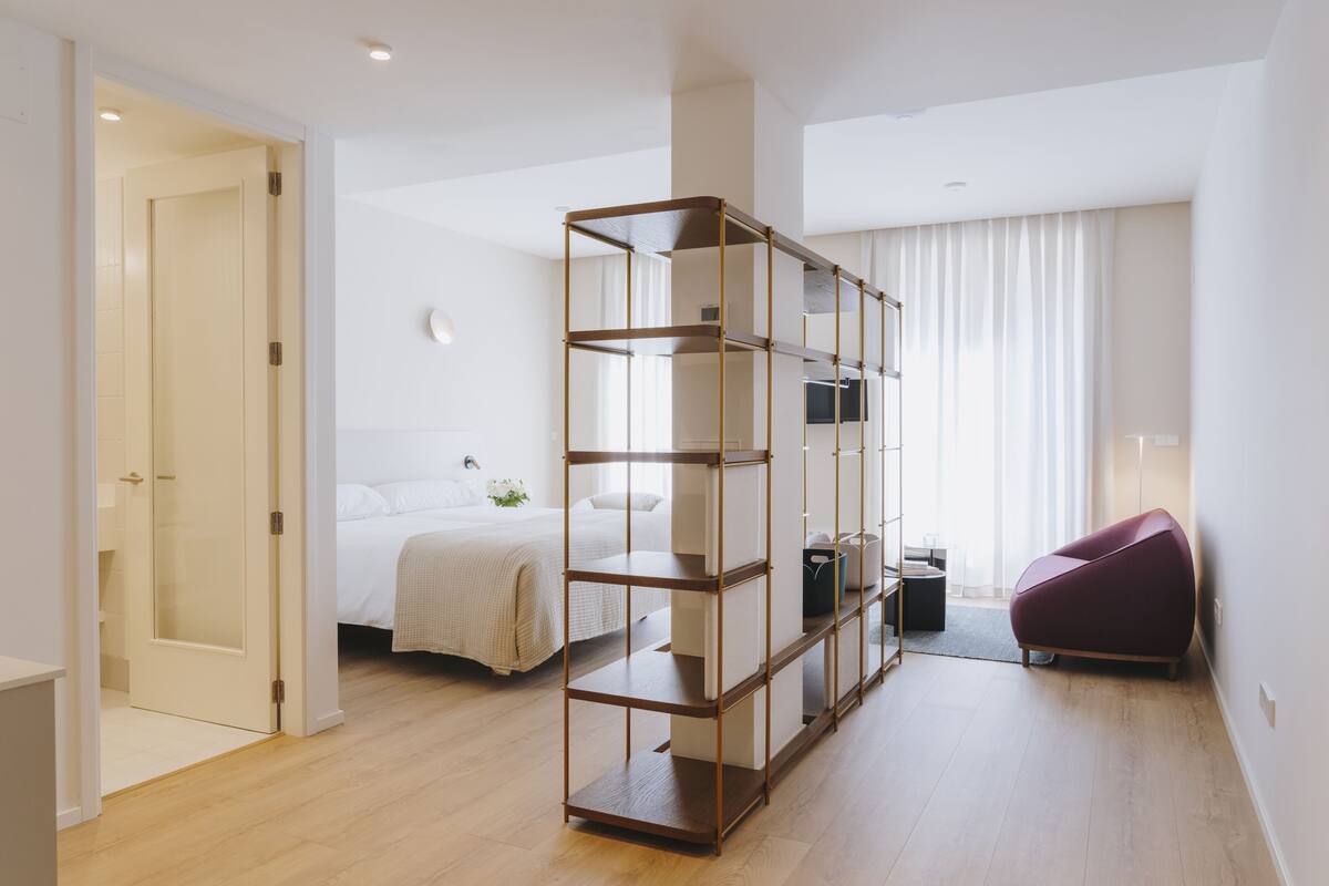 A modern hotel room with a wooden floor, a large bed, a built-in bookshelf, a seating area, and a door leading to a bathroom.