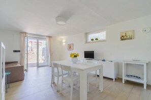 Um quarto claro e branco com uma mesa de jantar, cadeiras, uma televisão e uma janela com cortinas.