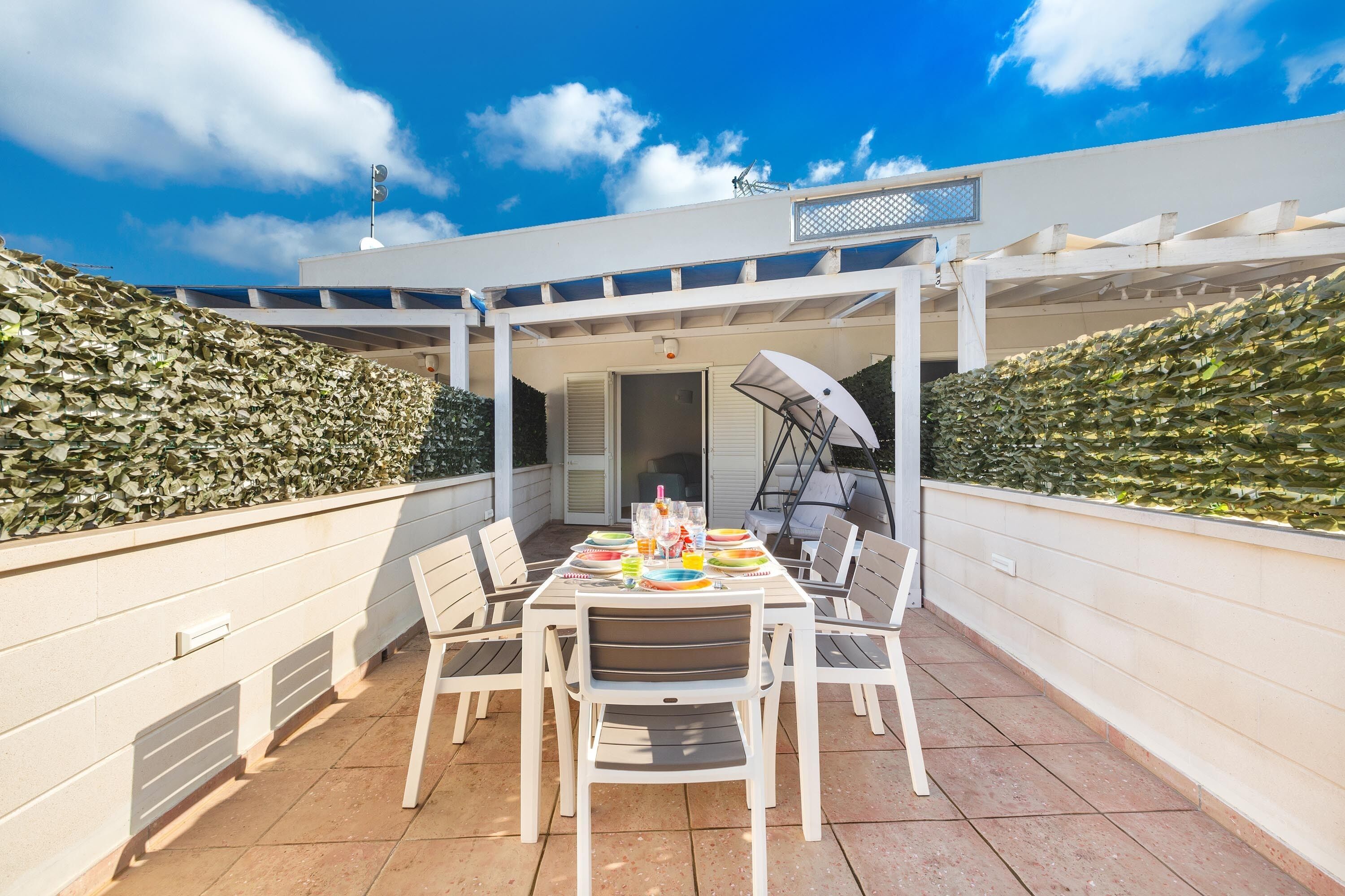 En terrasse med et bord dækket til et måltid, omgivet af stole, under et espalier.