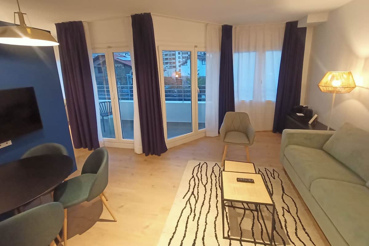 Een moderne woonkamer met een bank, fauteuils, een salontafel en een televisie. Grote ramen met gordijnen zijn zichtbaar, en er is een balkon te zien door de glazen deuren.