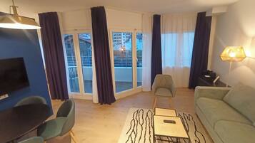 Un salón moderno con un sofá, sillas, una mesa de centro y un televisor. Se ven grandes ventanales con cortinas, y un balcón se aprecia a través de las puertas de cristal.