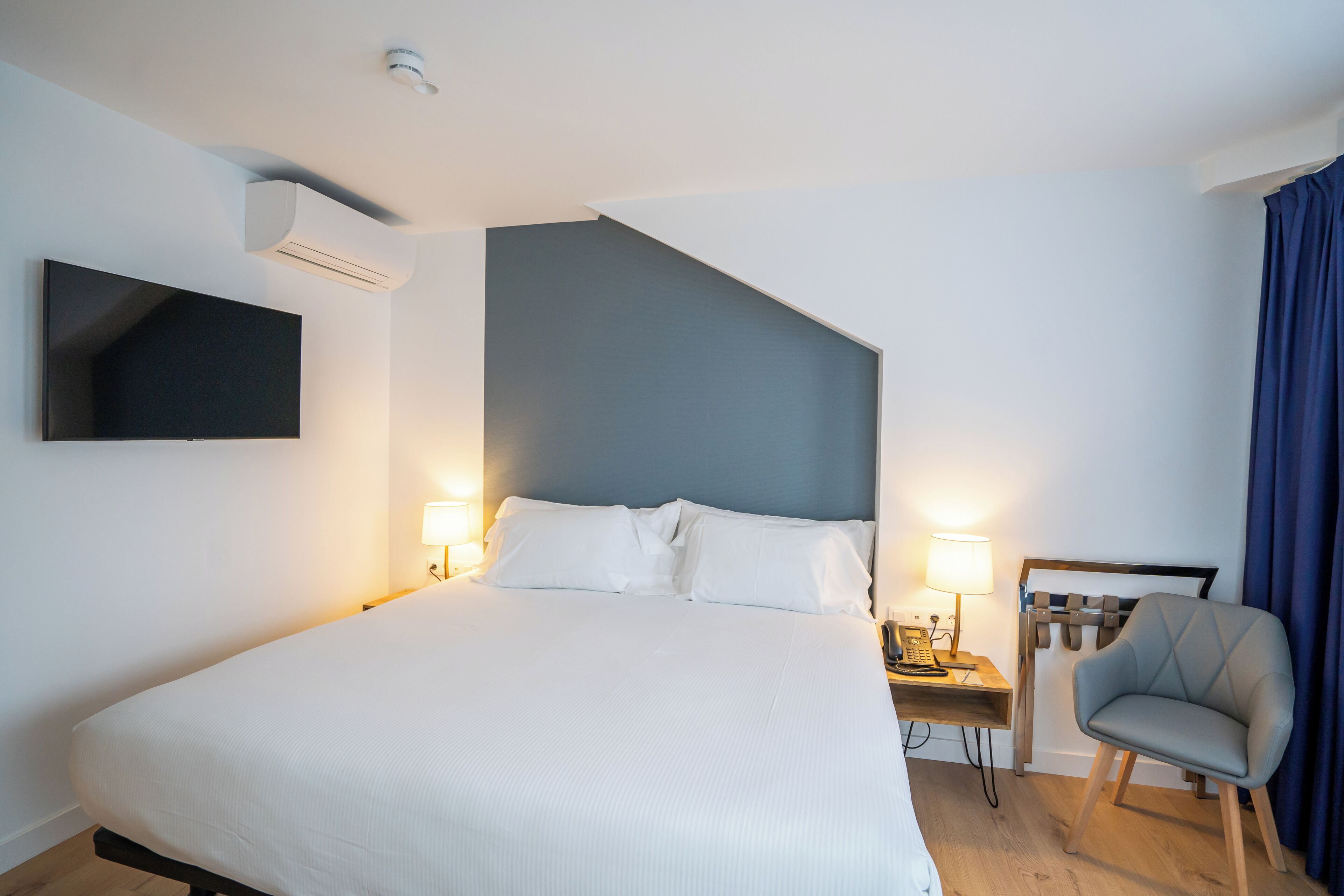 Una habitación de hotel moderna con una cama grande, lámparas de noche, un televisor de pantalla plana montado en la pared, un escritorio con una silla y una cortina azul.