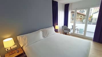Habitación de hotel con una cama grande, una mesita de noche con lámpara, una silla y vistas a edificios a través de la ventana.