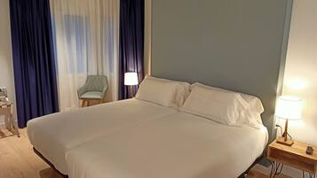 A large bed with white linens, a bedside table with a lamp, a chair, and a window with blue curtains.