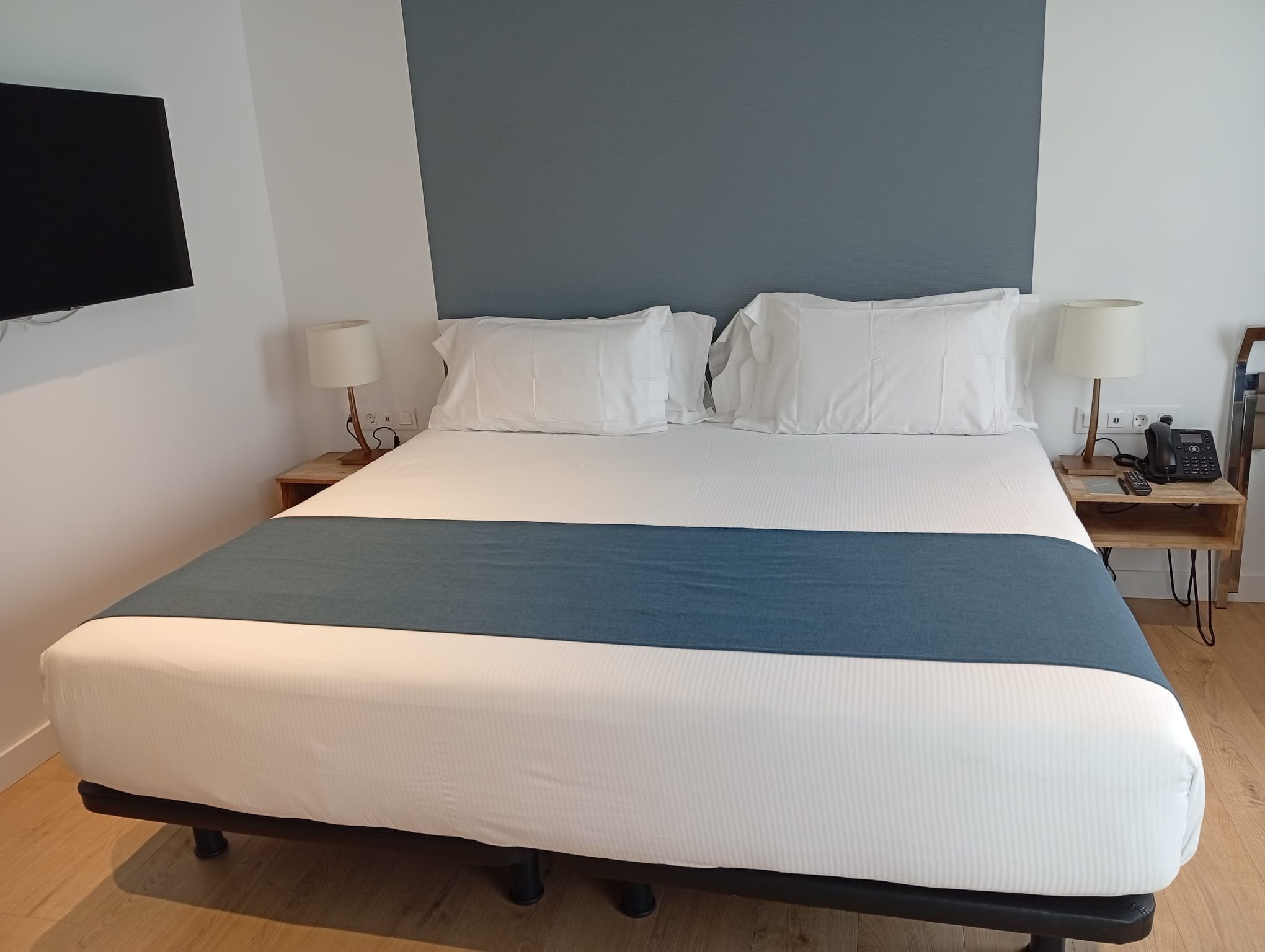 A large bed with a blue and white bedspread, two bedside tables with lamps, a phone, and a flat-screen TV mounted on the wall.